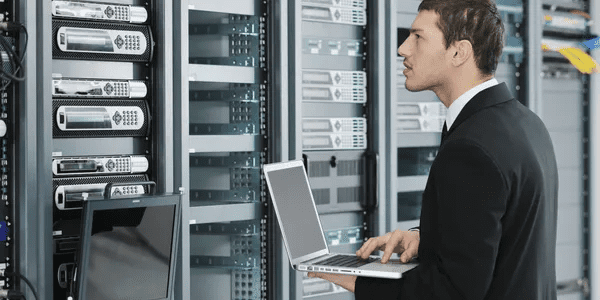 A man in black shirt holding laptop near server racks.